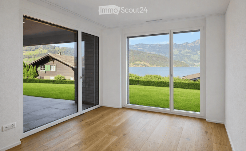 Empty room with wooden flooring, large windows, and sliding glass doors opening to a garden and scenic mountain view.