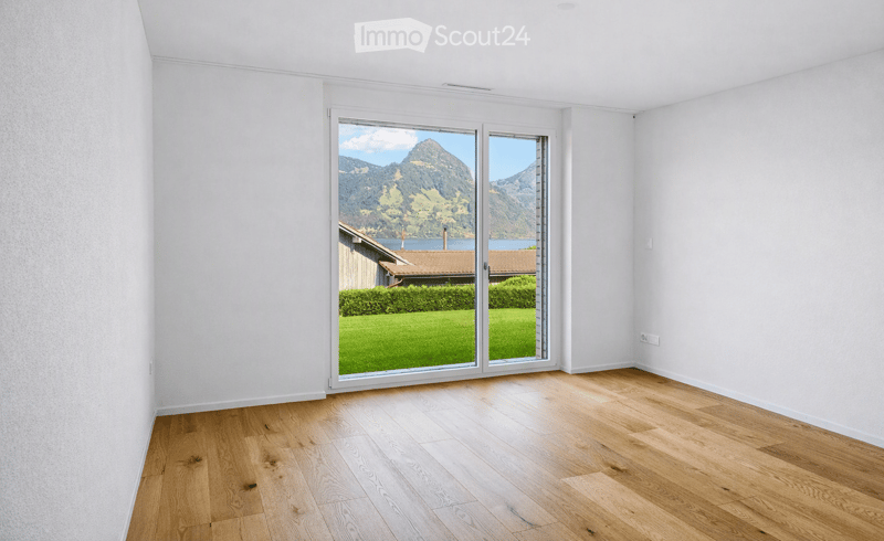 Empty room with light wooden flooring and white walls, featuring large sliding glass doors that open to a scenic view of mountains and a green lawn.