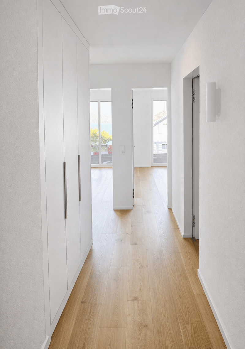 Modern hallway with light wooden flooring, white walls, built-in white cabinets, and open doorways leading to other rooms.