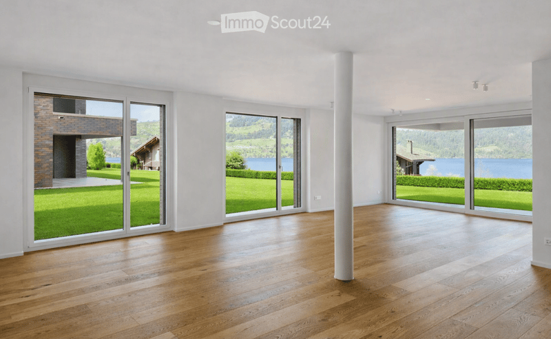Bright, empty room with wooden flooring, large glass sliding doors, and white walls. The room has a central white column and offers views of a green lawn and a lake.