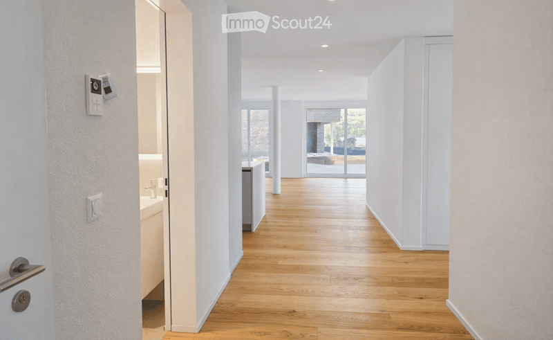 Bright, minimalist hallway with light wooden flooring, white walls, and recessed ceiling lighting. An open doorway leads to a room with a visible sink and faucet.