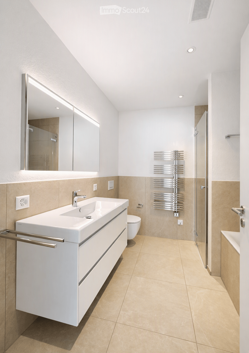 Modern bathroom with a white sink, chrome faucet, large mirror, radiator, and toilet. Light beige tiles on walls and floor, sleek design.