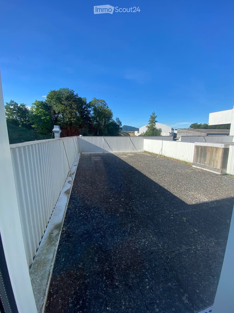 A narrow, gravel-covered balcony or terrace area enclosed by white metal railings, with a clear view of surrounding greenery and distant buildings.