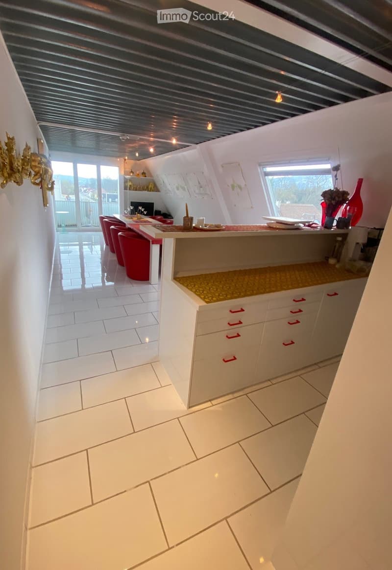 Modern kitchen with white cabinets, red stools, and a central island. Features a large window with a view, decorative vases, and a shelf with items.