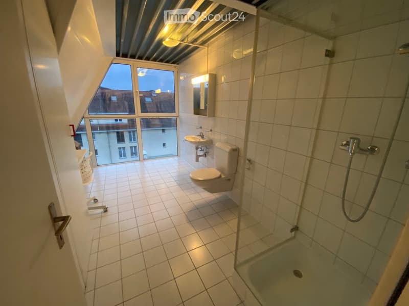 Modern bathroom with white tiled walls and floor, featuring a toilet, sink, mirror, and a glass-enclosed shower. Large windows provide natural light and a view of the city.