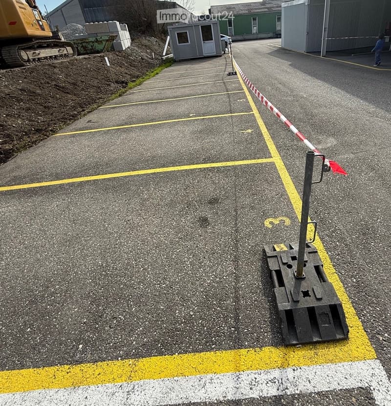 Paved outdoor area with marked parking spaces, yellow lines, and a temporary barrier. Construction equipment and a small portable building are visible in the background.