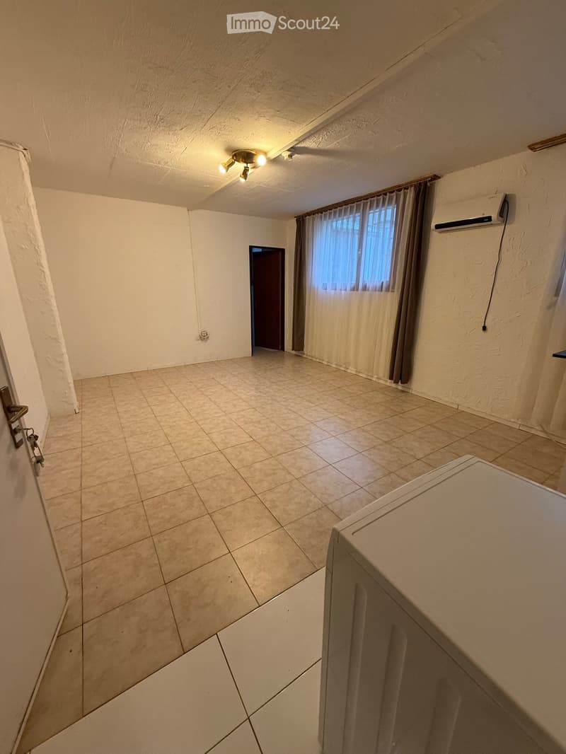 an empty room with tiled floor, white walls, a ceiling with lights, a window with white and brown curtains, a door, and an air conditioner on the wall
