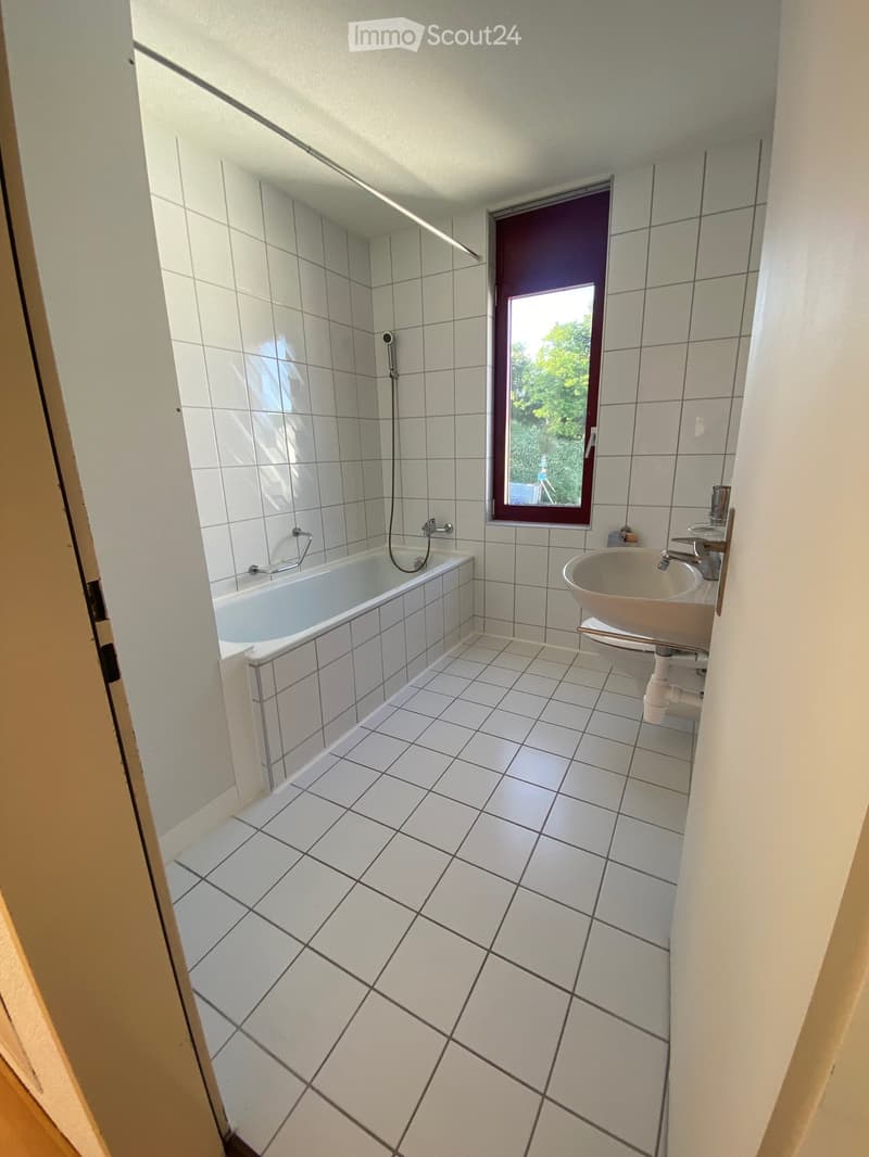 Modern bathroom with white tiled walls and floor, featuring a bathtub, sink, and toilet. A window with a purple frame provides natural light.