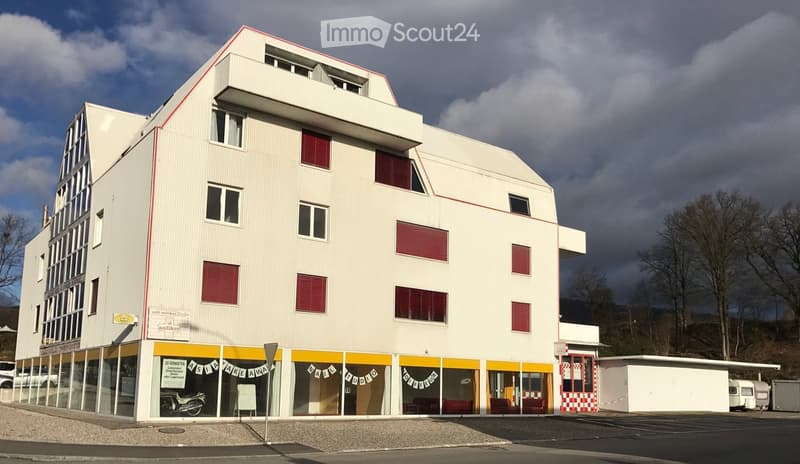 Modern multi-story building with white facade, red shutters, and storefronts including a nail studio and take-away restaurant. The building has a contemporary design with large windows and a flat roof.