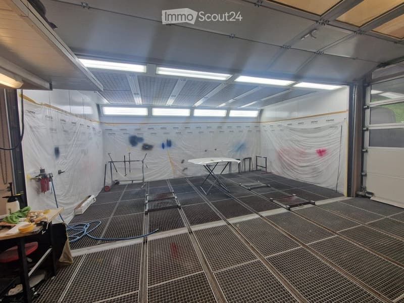 Empty room with metal grated flooring, white walls covered in protective sheets, and minimal equipment including a small table and stools.