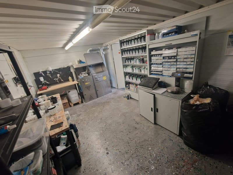 The image shows a cluttered workshop or storage area with various tools, shelves filled with paint cans, a workbench, and miscellaneous items scattered around. The space has a concrete floor and metal shelving units.
