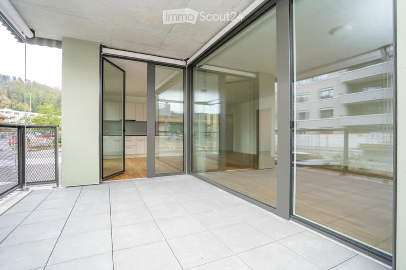 Modern balcony with large glass sliding doors, white tiled floor, view of greenery and neighboring buildings.
