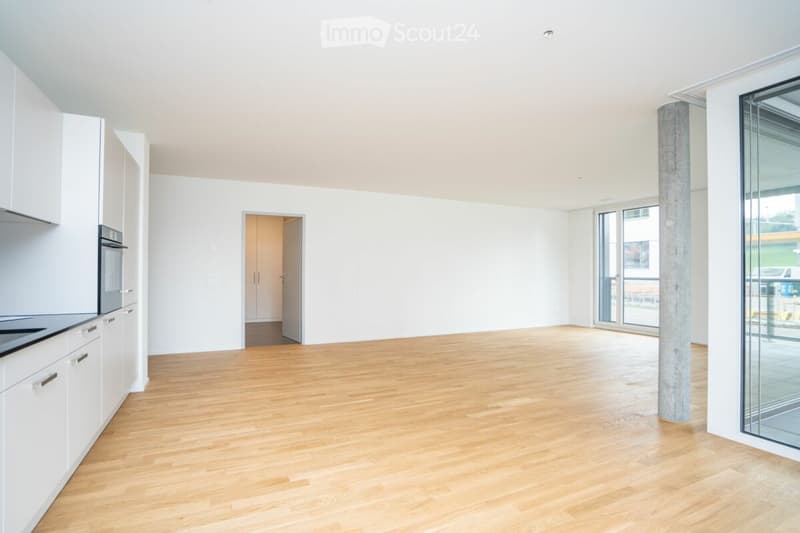 Open-plan unfurnished room with light wooden flooring, white walls, and modern white kitchen units on one side. Features large glass sliding doors leading to a balcony area and a doorway to another room.