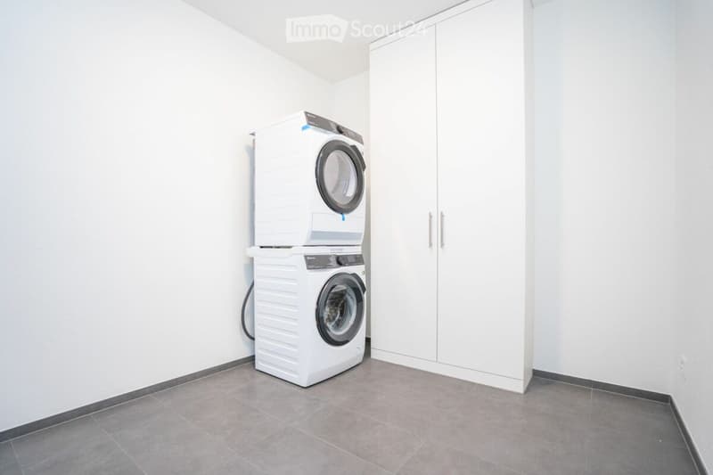 Modern, minimalist laundry room with white walls, grey tiled floor, and built-in white cabinetry. Features a stacked washer and dryer unit.