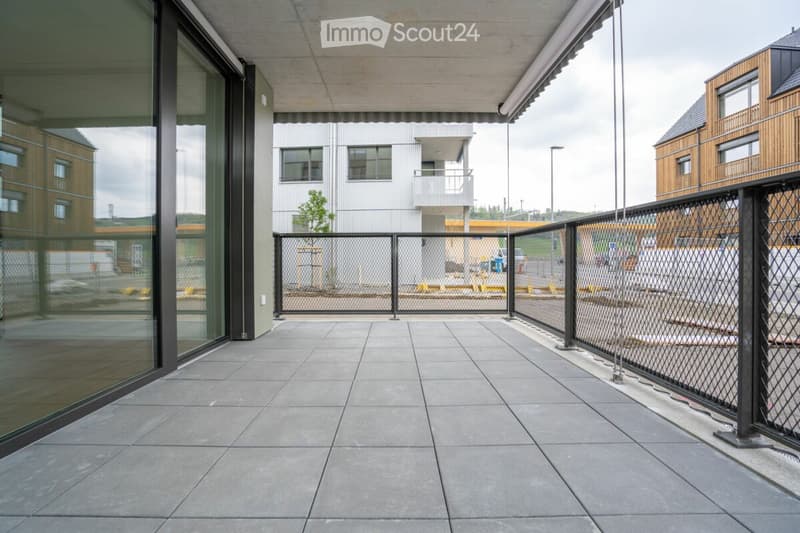 Modern balcony with tiled floor, glass walls, and metal fencing, overlooking a secured area with other buildings in the background.