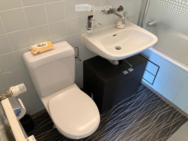Modern bathroom with a white toilet, sink, and dark cabinet. The floor has a black and white abstract pattern, and the walls are tiled in light grey.