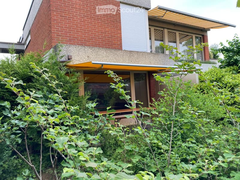 A modern house with red brick walls, large windows, and a yellow awning, partially obscured by lush green bushes and trees.