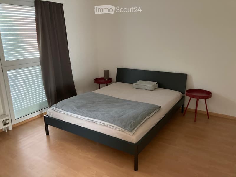 A modern bedroom with a single bed, a nightstand, a small table, and a lamp. The room features wooden flooring and large windows with blinds.