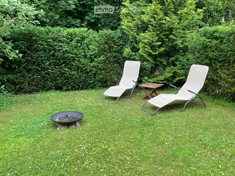 Garden with two white sun loungers, a small wooden table, and a black fire pit on a grassy lawn surrounded by dense green bushes.