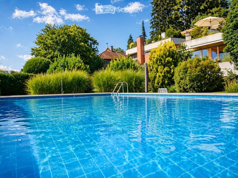 A swimming pool surrounded by greenery, with a modern building and umbrella in the background.