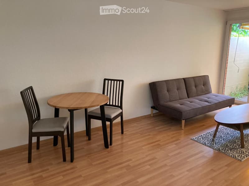 A small, modern living room with a sofa, a round wooden table, and two chairs. The room features wooden flooring and a patterned rug.