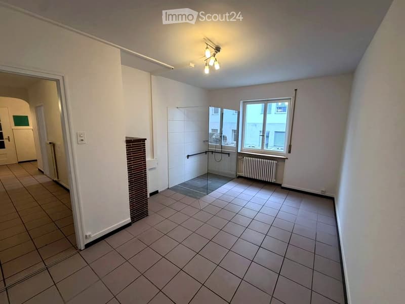 Empty room with tiled floor, white walls, and a glass-enclosed shower area visible in the background. The room has a window with a radiator below it and a door leading to another area.