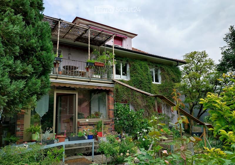 A two-story house with a balcony and a well-maintained garden. The house features a brick facade, large windows, and a covered entrance. The garden includes various plants, potted flowers, and a bench.