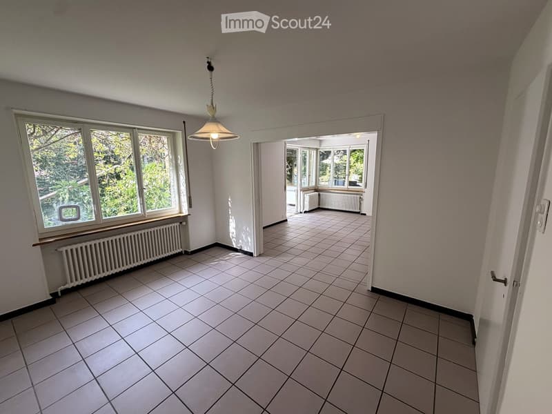 Empty room with tiled floor, white walls, large windows, and a hanging light fixture. Radiators are visible beneath the windows.