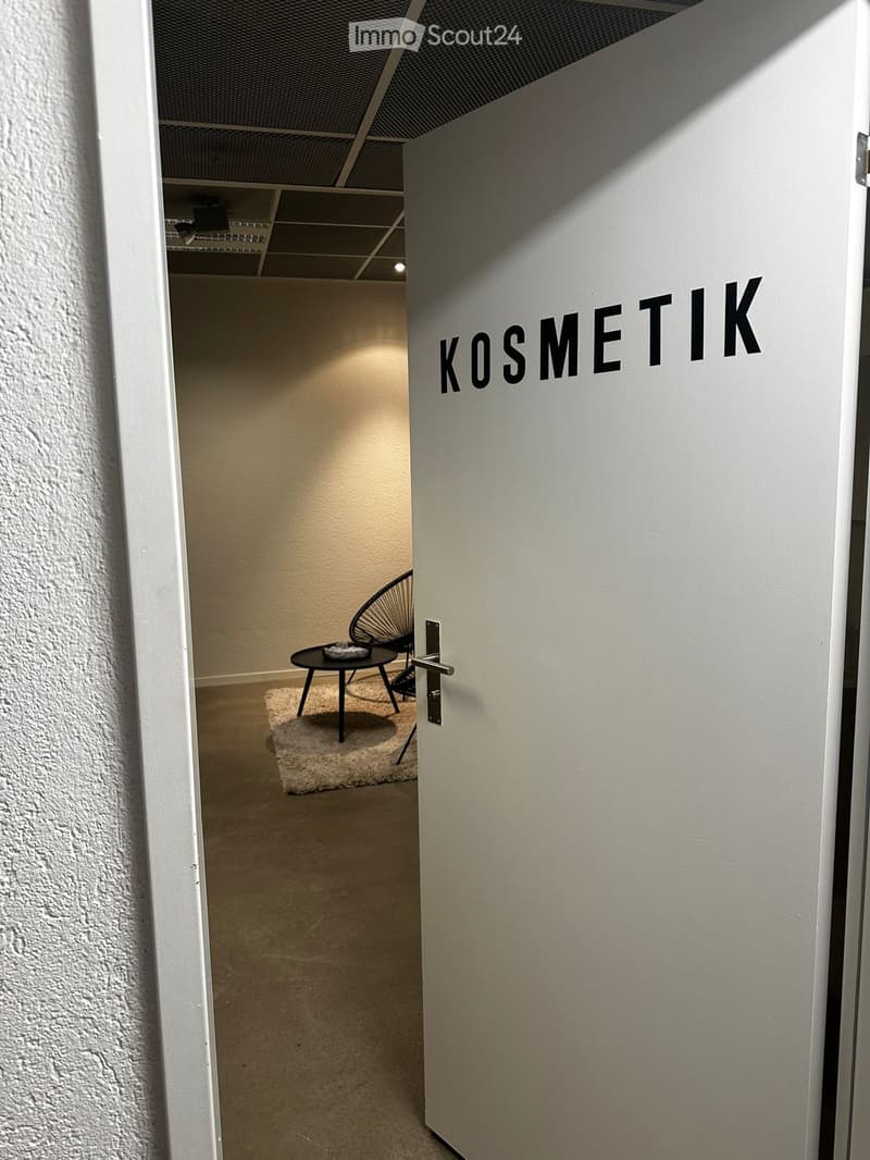 A small, furnished room with a white door labeled "KOSMETIK" and a visible interior containing a black wire chair, a small black table, and a white rug.