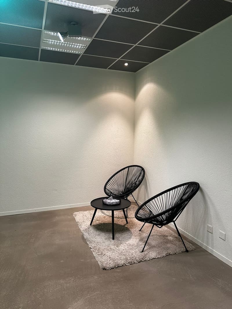 Corner of a room with two black wire chairs, a small black table, and a white rug on a concrete floor.