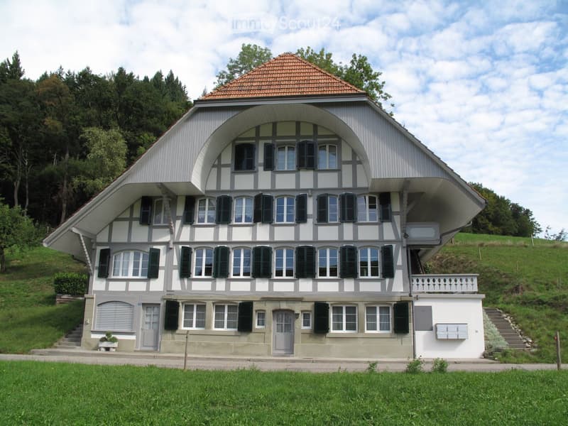 Two-story house with a distinctive curved roof and multiple windows, surrounded by greenery and a grassy lawn.
