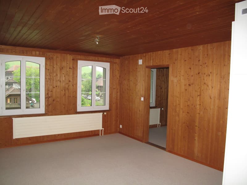 Empty room with wooden walls and ceiling, carpeted floor, two windows, radiator, and a doorway leading to another room.