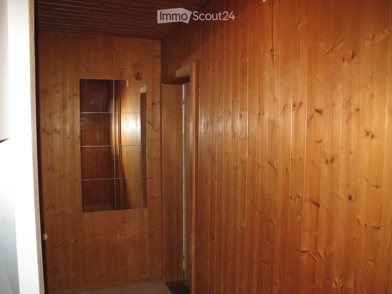 Wooden-paneled hallway with a mirrored door and a small shelf, featuring a wooden ceiling and light-colored wooden flooring.