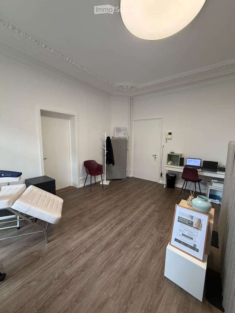 A modern, well-lit room with wooden flooring, white walls, and a decorative ceiling. Features include a white leather stool, a red chair, a small table with a vase and picture frame, a desk with a laptop and screen, and a coat rack with a jacket.
