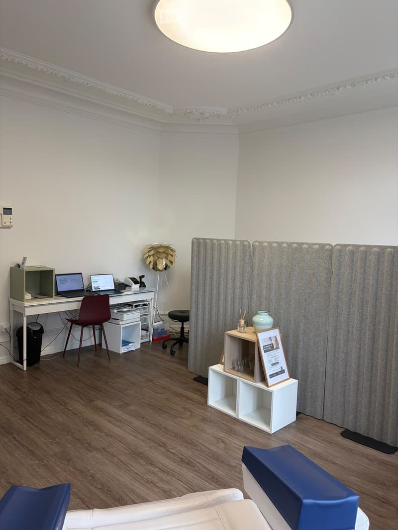 A modern, well-lit room with a wooden floor, white walls, and decorative lighting. It features a desk with a laptop, a red chair, a small table with a vase and cup, and a white couch with blue cushions.
