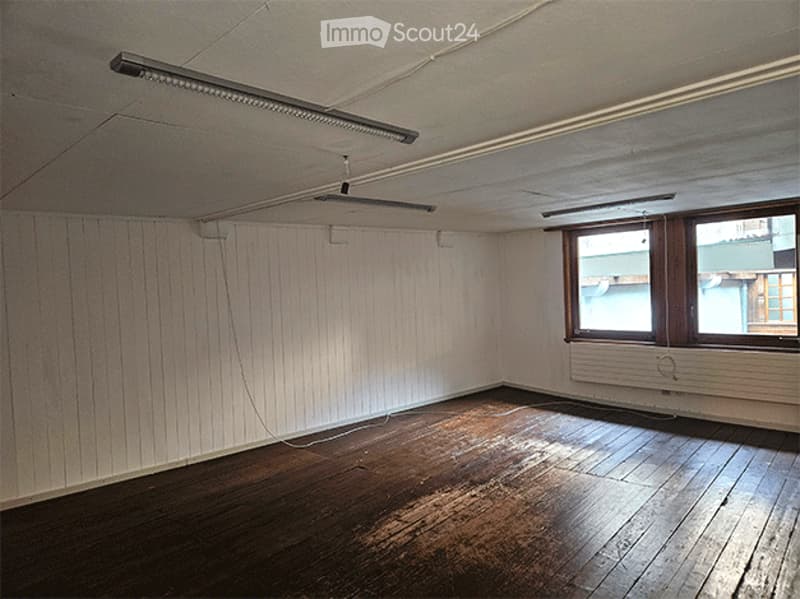 Empty room with wooden flooring, white paneled walls, and two windows with wooden frames. Exposed electrical wiring and a radiator are visible.