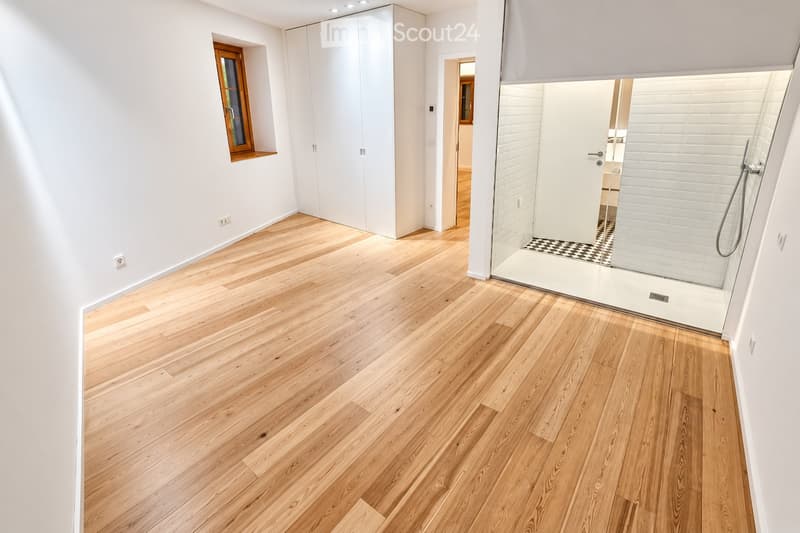 Empty room with light wooden flooring, white walls, built-in wardrobes, and a glass-enclosed shower area visible in the background.