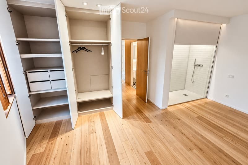Empty room with light wooden flooring, white walls, and an open closet with shelves and drawers. A glass-enclosed shower area is visible in the background.