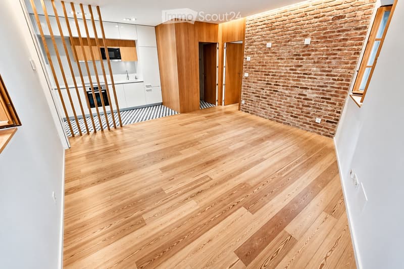 Empty room with light wooden flooring, white walls, and a brick accent wall. The room features a modern kitchen area with white cabinets and wooden accents visible in the background.