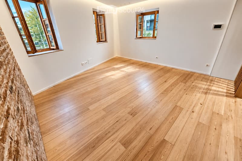 Empty room with light wooden flooring and white walls, featuring multiple windows with wooden frames and natural light streaming in.