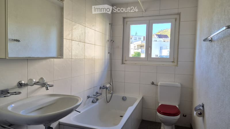 Bathroom with white tiles, a sink, a bathtub, a toilet with a red seat, a mirror cabinet, and a window offering a view of the outside.