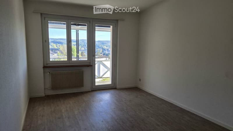Empty room with wooden flooring, large window with a view, radiator, and a sliding door leading to a balcony.