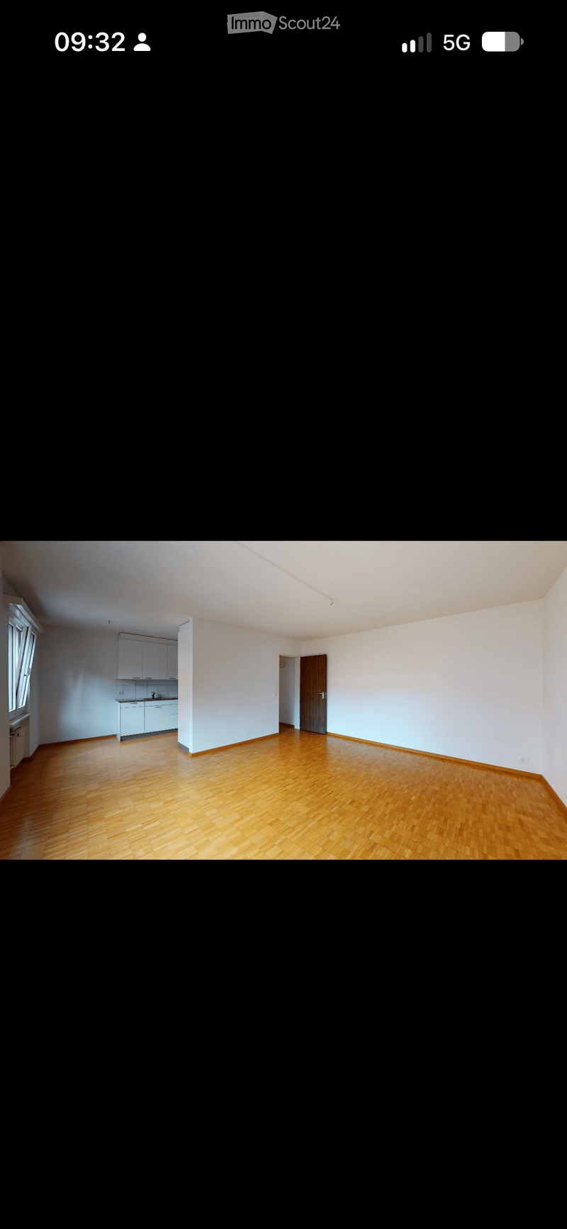 Empty room with wooden flooring, white walls, and a small kitchen area visible in the background.