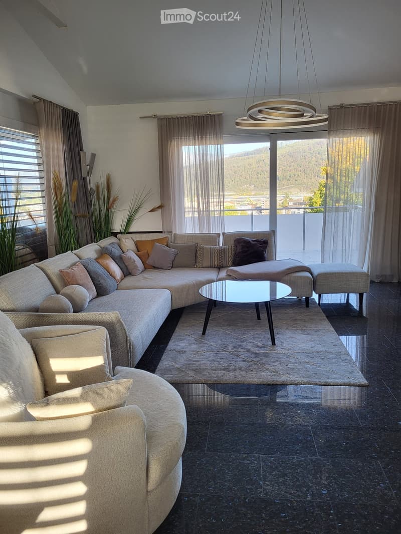Modern living room with a large sectional sofa, coffee table, ottoman, pendant light, and a view of the landscape through large windows.