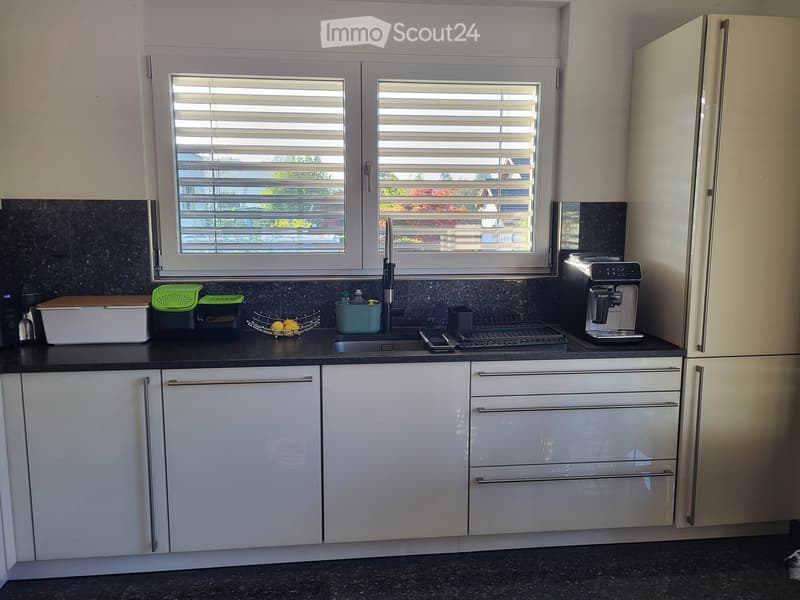 Modern kitchen with white cabinets, black granite countertop, stainless steel sink, and a window with blinds. Includes a refrigerator, coffee maker, and various small items on the counter.