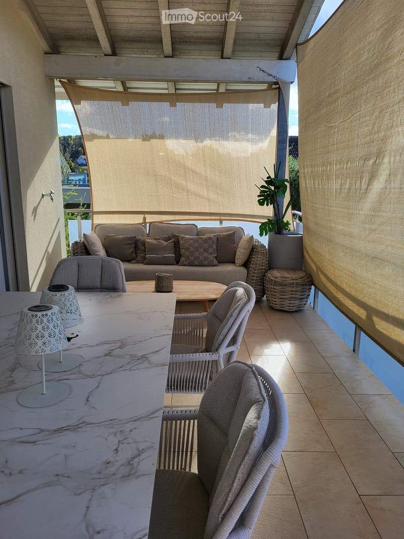 A modern outdoor balcony with a sofa, coffee table, chairs, lamps, and a potted plant, featuring a retractable shade and a marble countertop.