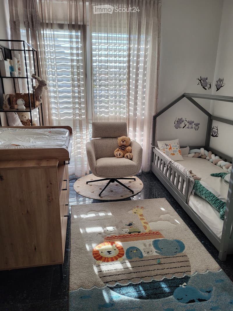 A cozy children's bedroom with a baby bed, a plush chair, a shelf with toys, and a colorful rug featuring animal prints.