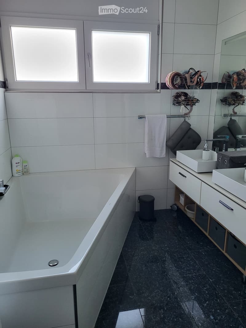Modern bathroom with a bathtub, double sink, large mirror, and white tiled walls. Includes a towel rack, storage cabinet, and toiletries.
