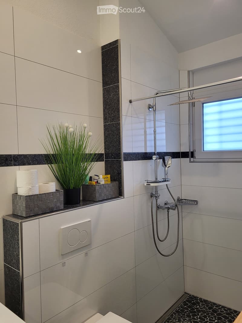 Modern bathroom with white tiles, a glass-enclosed shower, a sleek faucet, and a small cabinet with a decorative flower arrangement and toiletries.