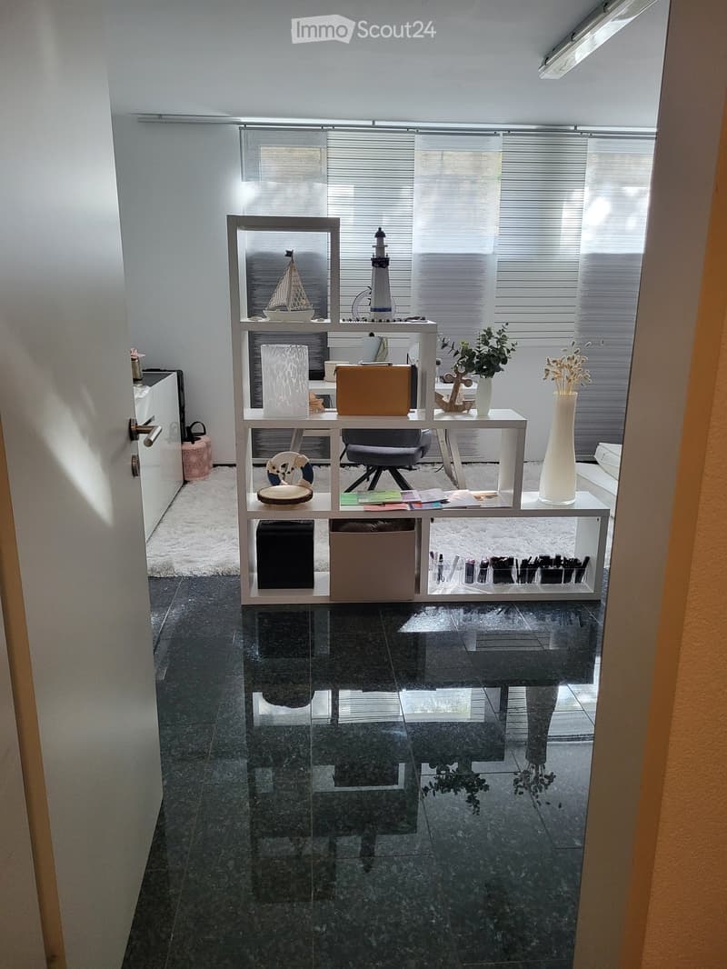 A modern, well-lit room with a sleek black tiled floor, a white shelving unit holding various decorative items, a chair, and a small table with a vase and books.